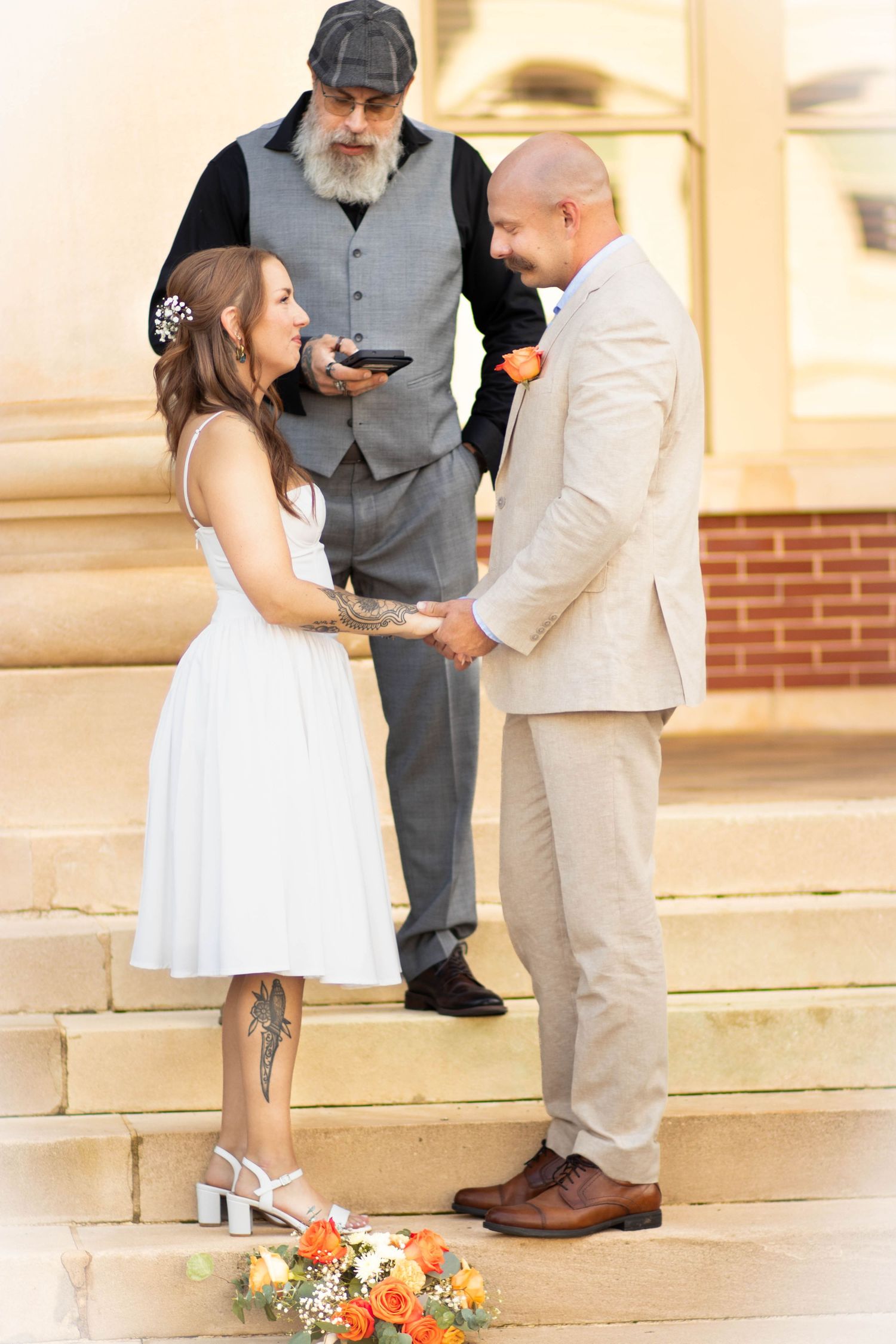 Bride and groom hold hands on stone steps during outdoor wedding ceremony officiated by bearded man in gray vest.