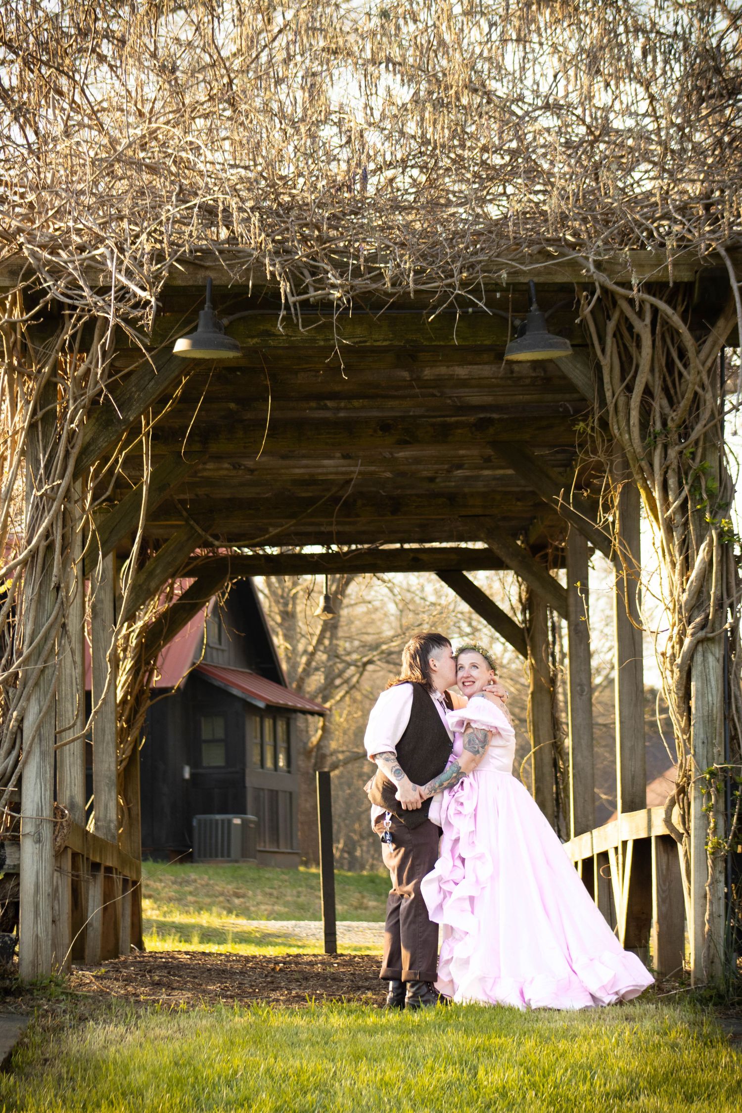 A couple in wedding attire embraces under a rustic wooden arbor covered in bare vines.