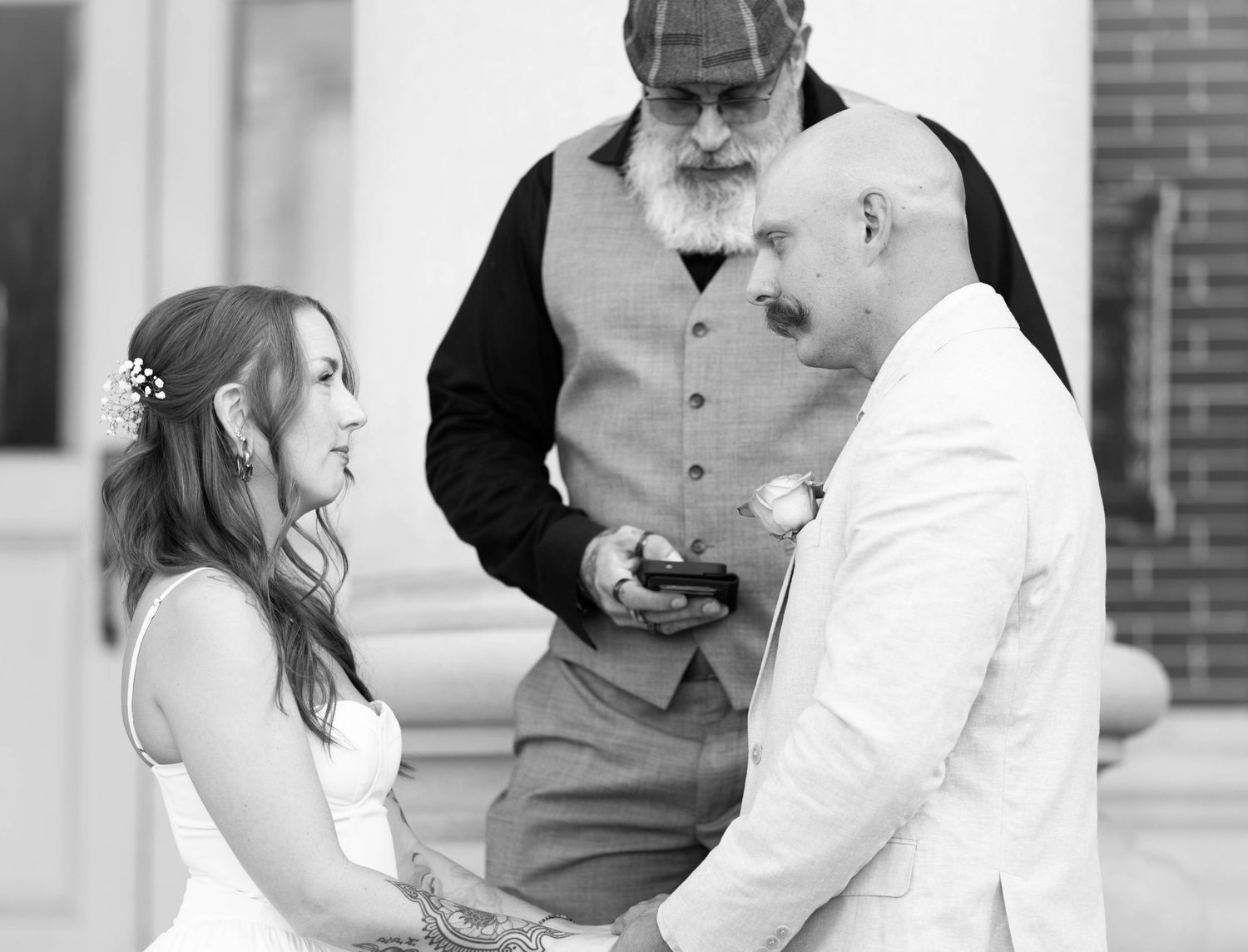 Black and white photo of bride and groom exchanging vows with bearded officiant during outdoor wedding ceremony.