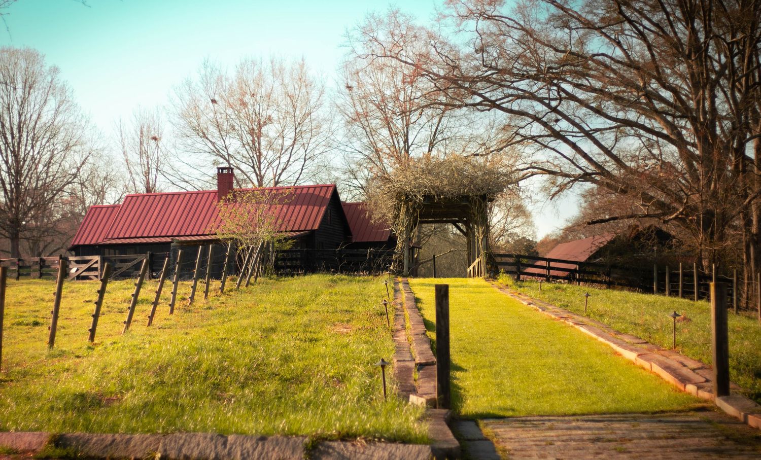 A rustic red-roofed barn surrounded by lush green grass, wooden fences, and bare trees on a sunny farm day.