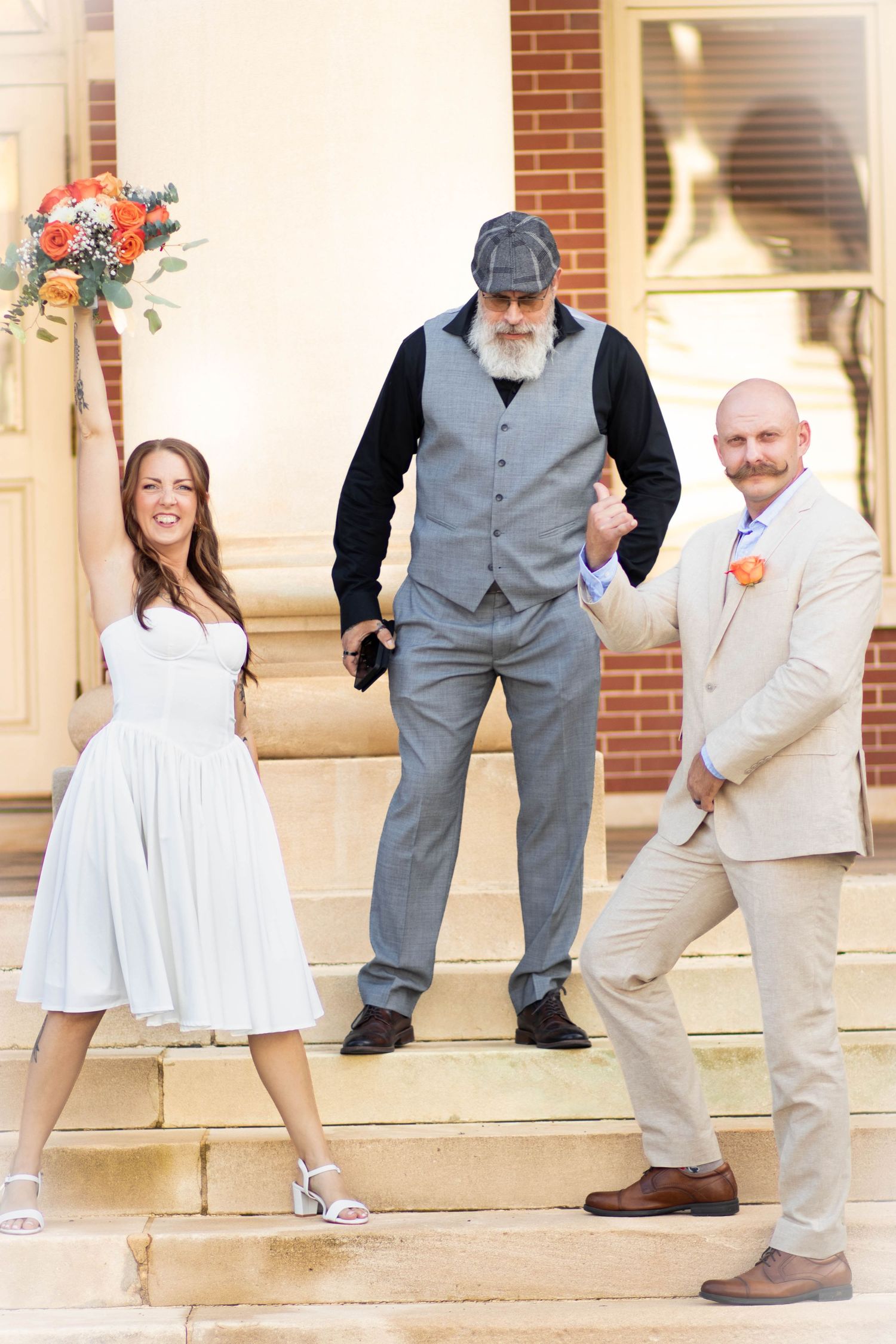 Bride in white dress holds bouquet aloft beside groom in tan suit, officiant in gray vest on stone steps.