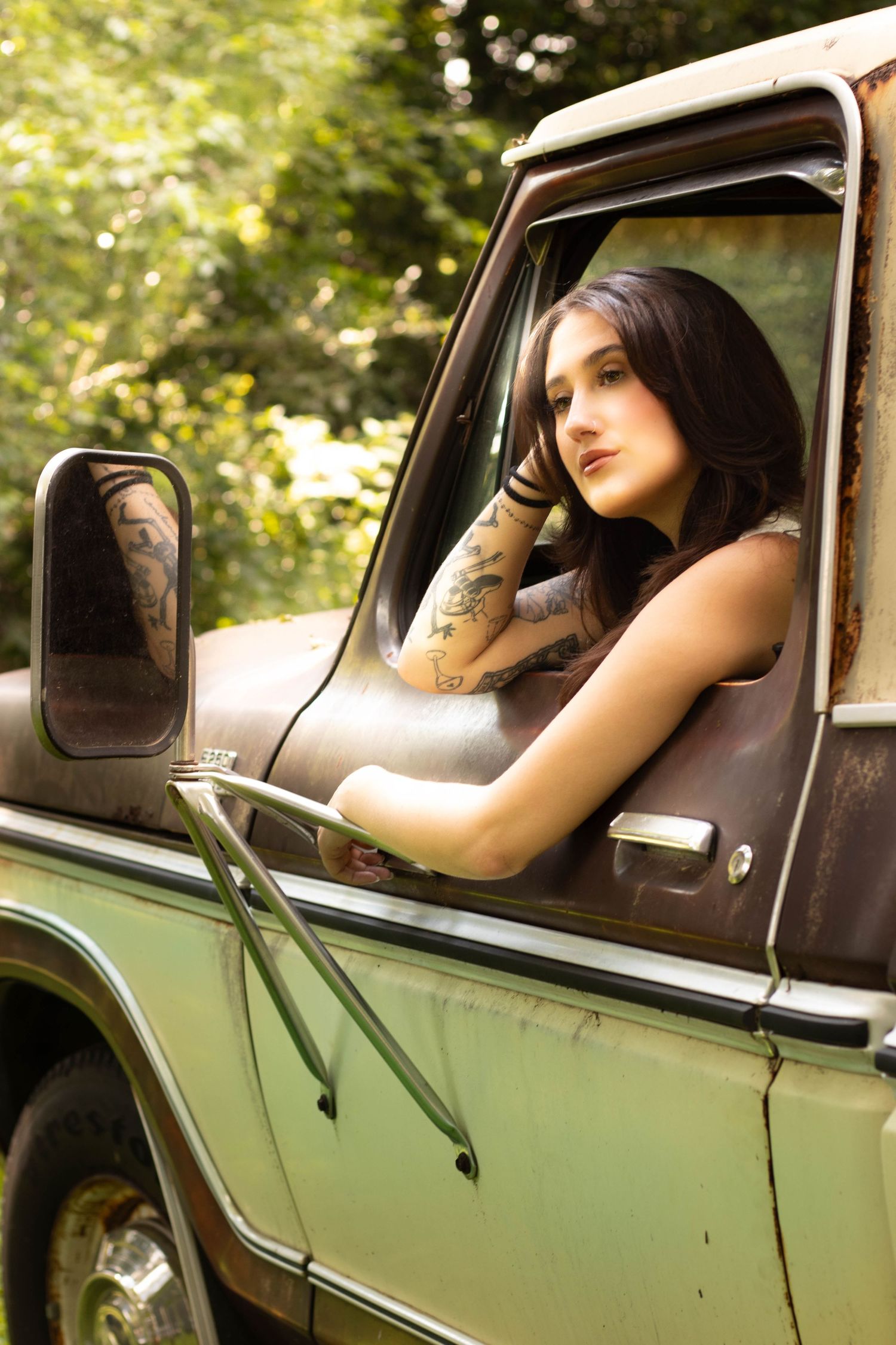 Tattooed young woman with dark hair leaning out window of vintage rusty green truck surrounded by trees.