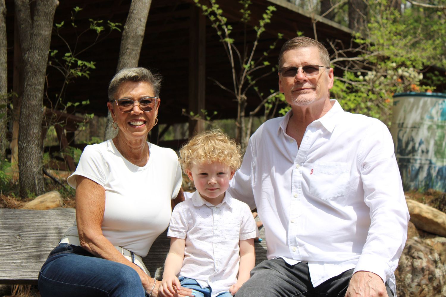 Grandparents sitting outdoors with young grandson on bench in wooded park setting.