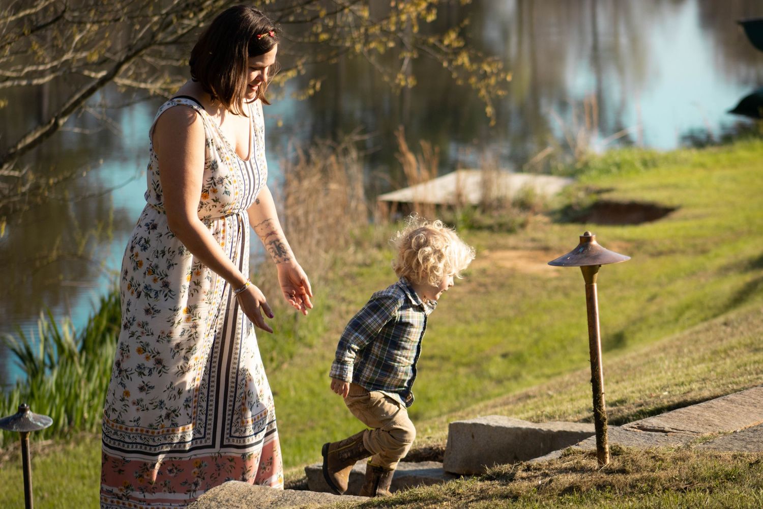 A mother in a floral dress walks with her toddler boy near a sunlit pond in a lush green garden.