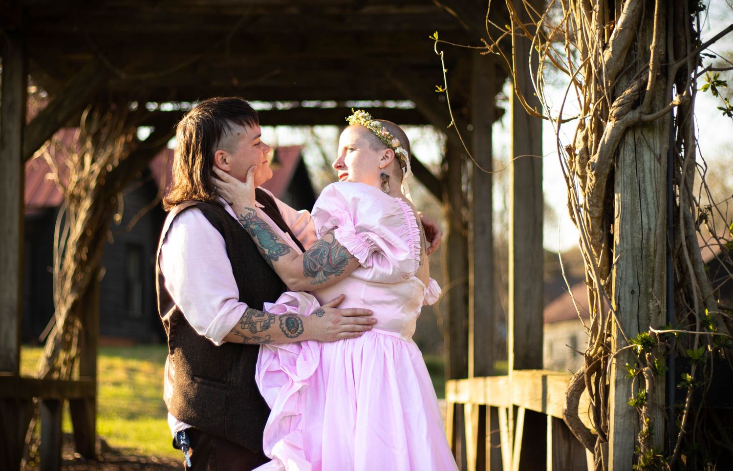 A tattooed man holds a young girl in a pink princess dress outdoors near a rustic wooden structure.