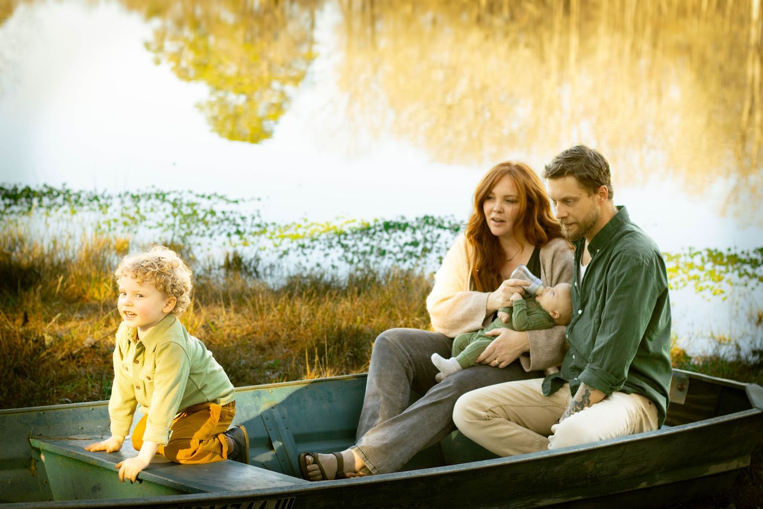 Family of three sitting in a green rowboat by a scenic pond, parents holding baby while toddler sits nearby.