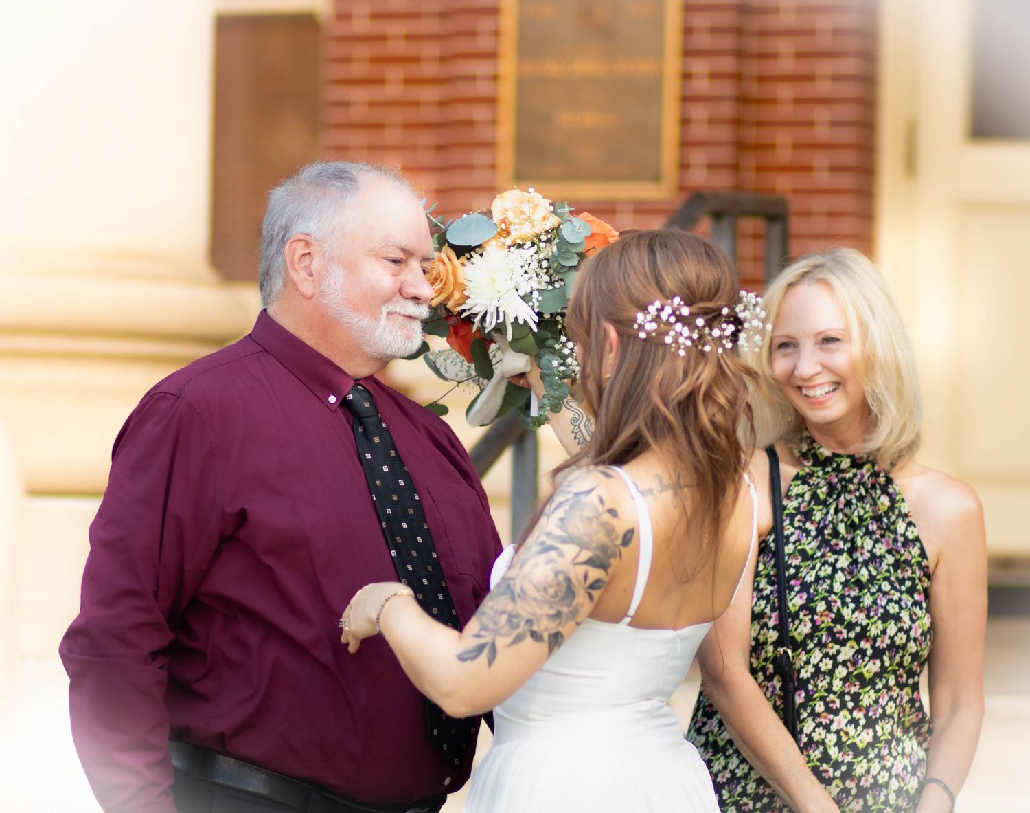 Bride with tattoos holding bouquet stands with older man in burgundy shirt and smiling blonde woman at outdoor wedding.