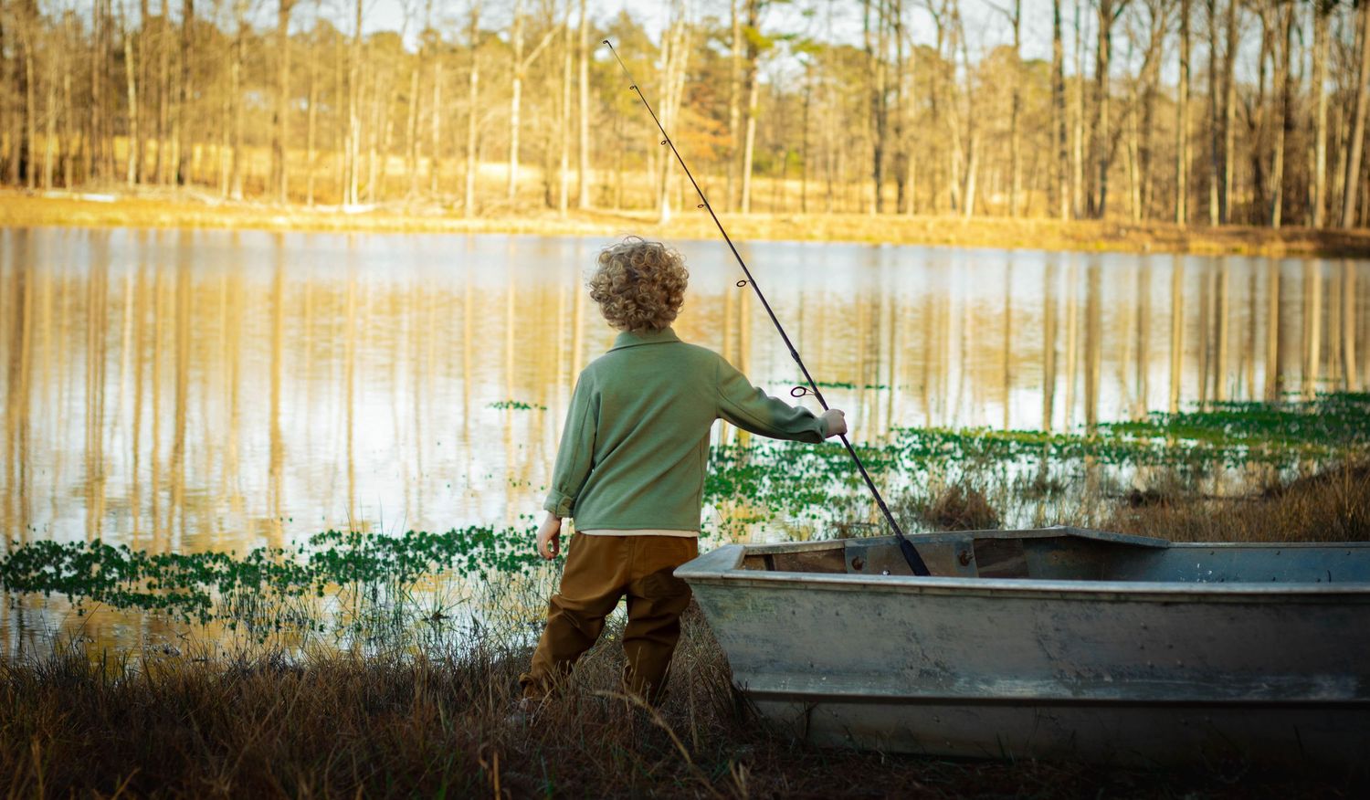 Young boy fishing by a calm lake with a rowboat, surrounded by lush greenery and tall trees in warm light.