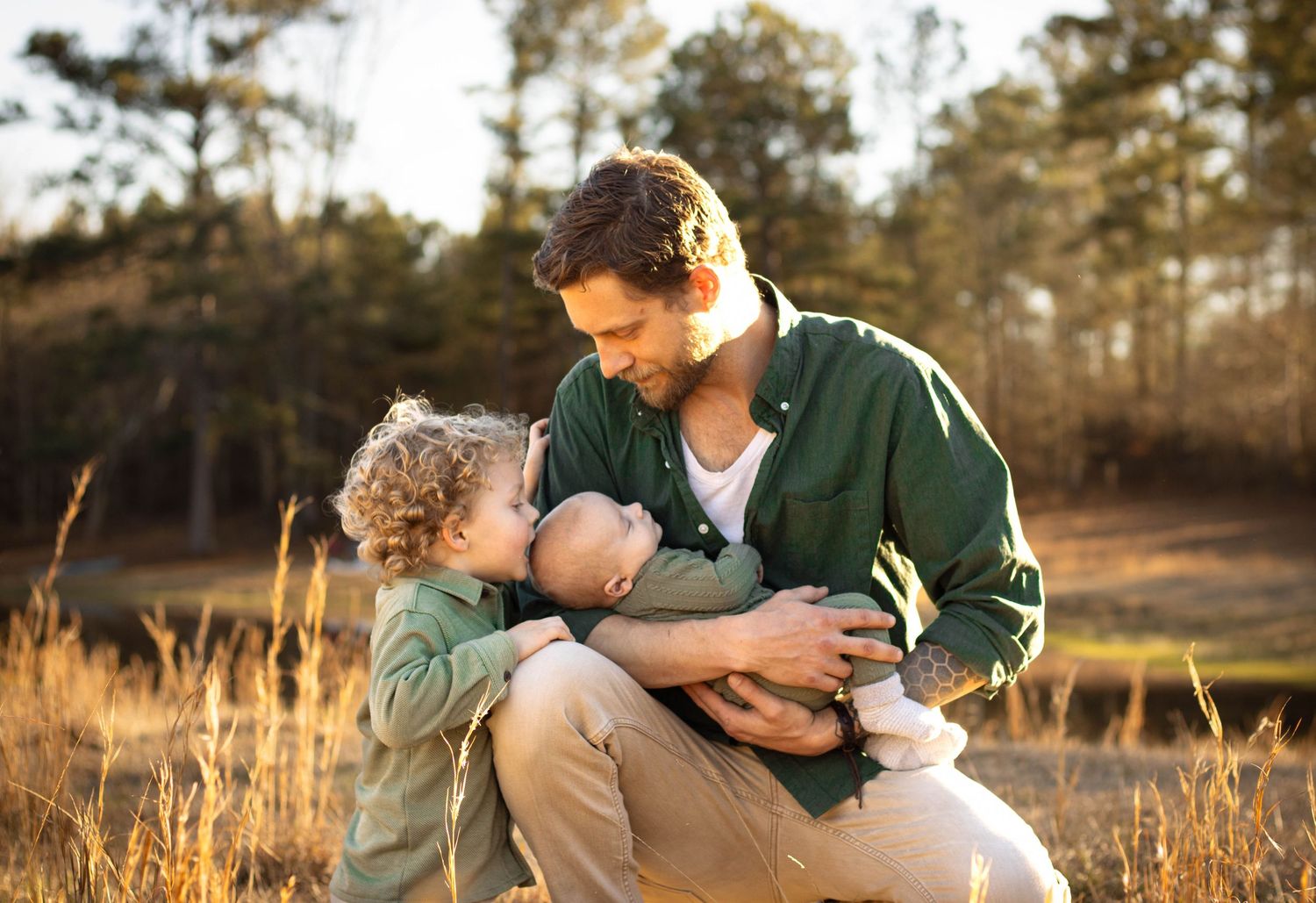 A father sits in a golden field holding his baby while his young curly-haired child leans in to give the infant a kiss.