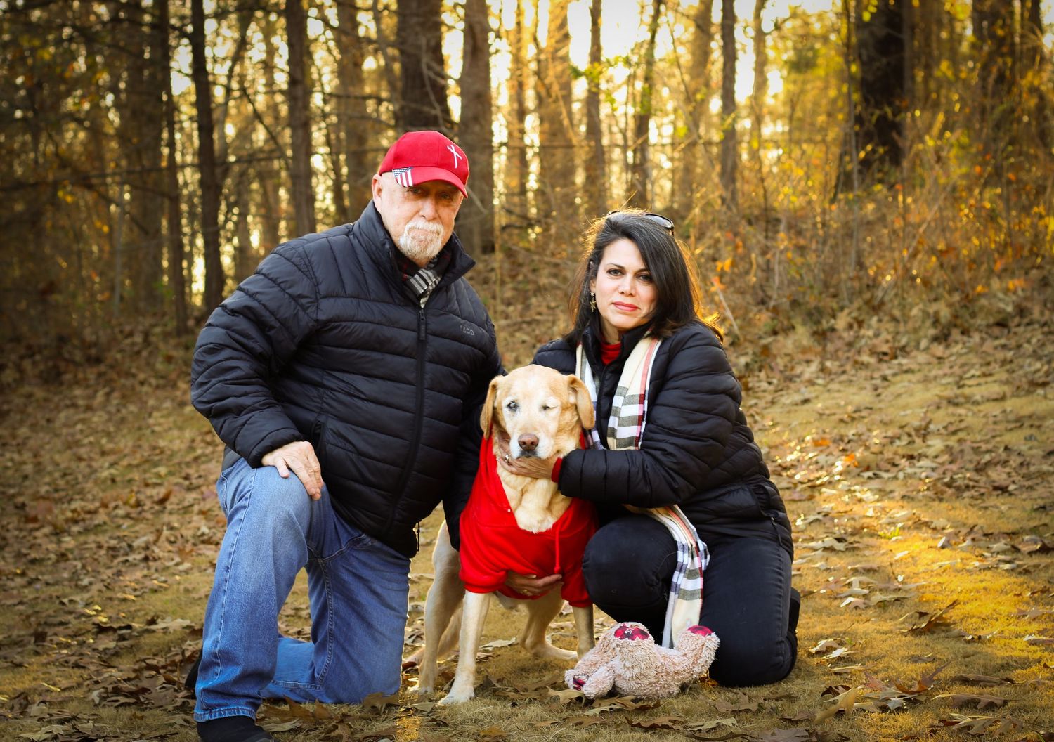 Man and woman kneeling with golden retriever in red outfit among fall foliage in woods.