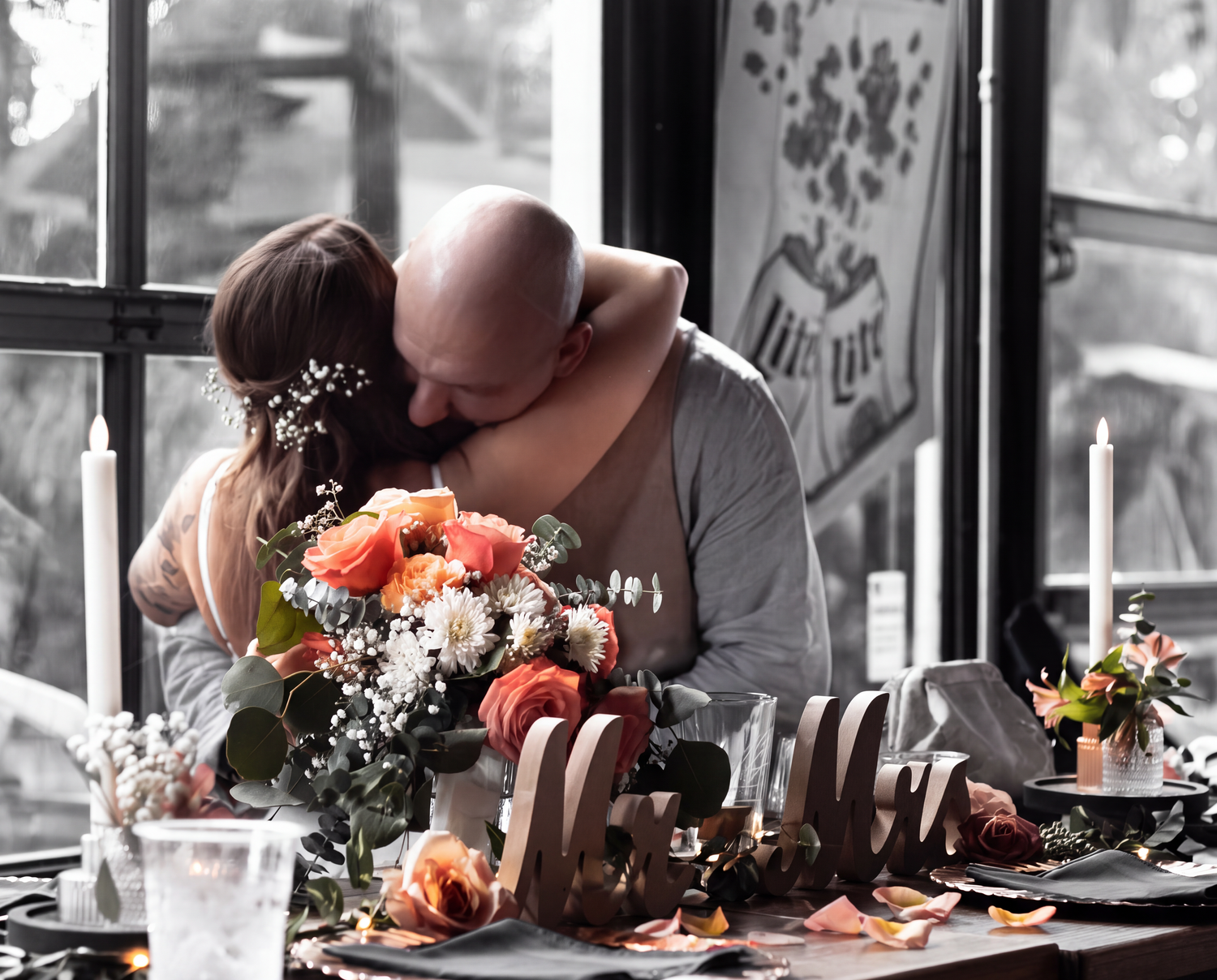 Couple sharing a romantic embrace at a decorated table with peach roses and candles in a rustic venue.