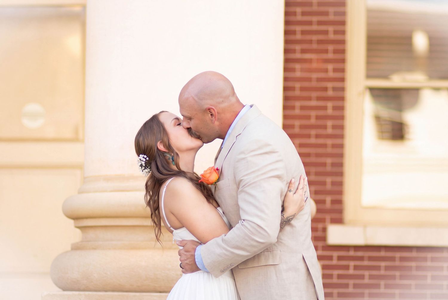 Bride and groom sharing a romantic kiss outdoors near brick building columns on their wedding day.