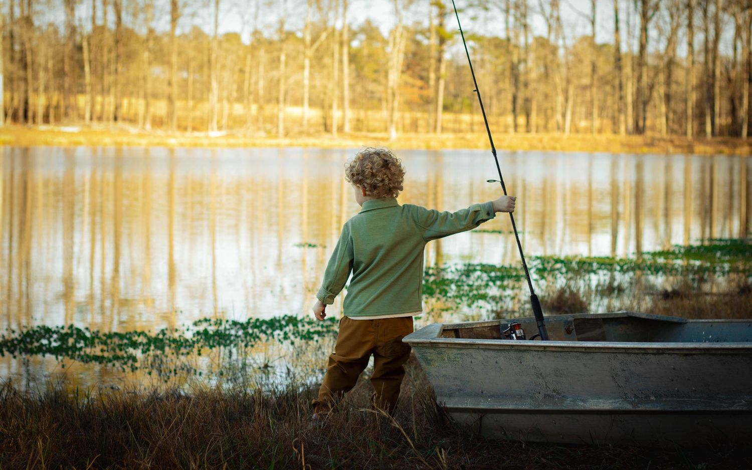 Young boy fishing with a rod by a calm lake next to a rowboat, surrounded by golden autumn trees.