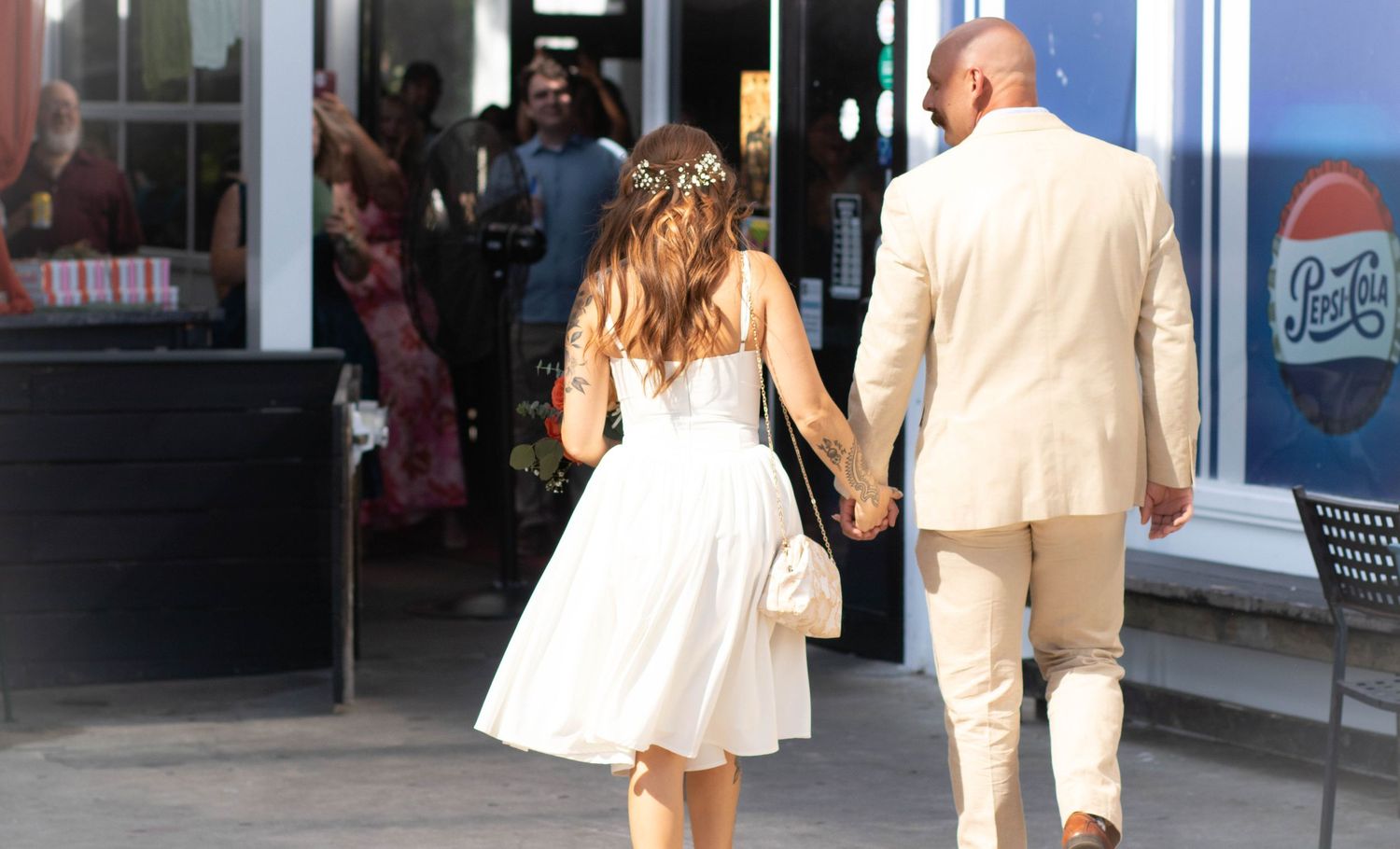 Newlywed couple holding hands walking toward a restaurant entrance, bride in white dress and groom in beige suit.