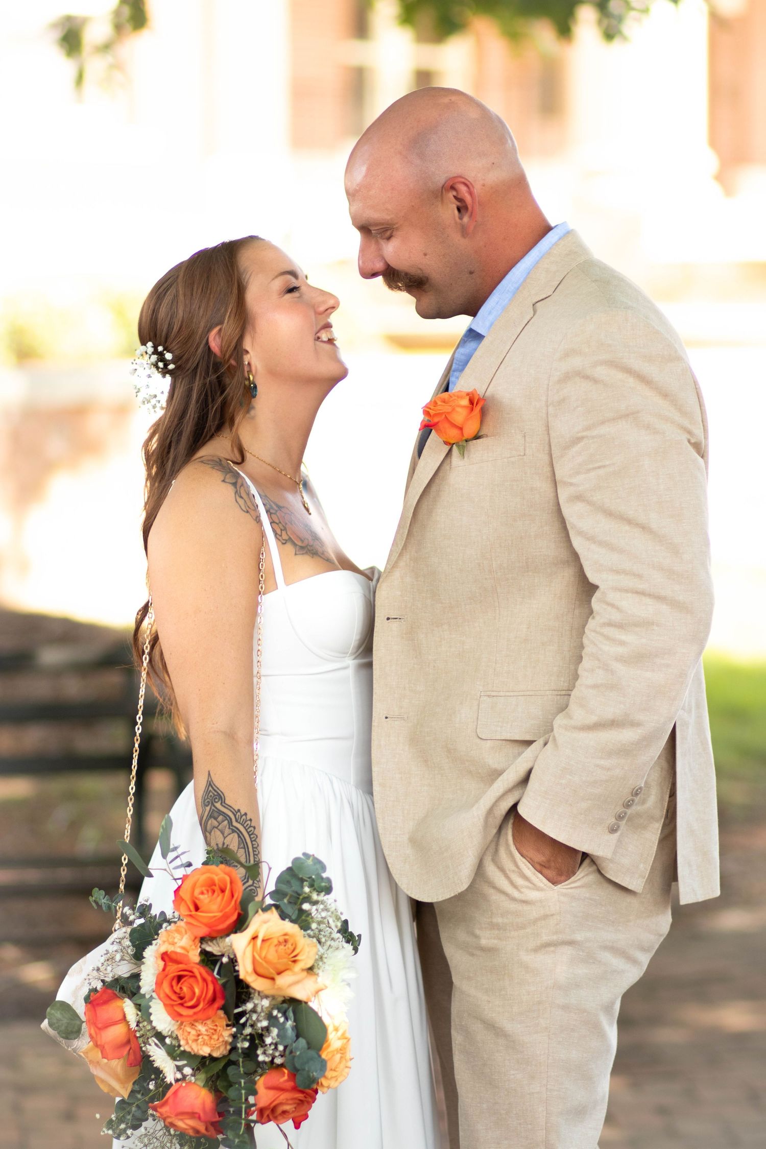 Smiling bride in white dress holding orange roses bouquet gazes at bald groom in tan suit at outdoor wedding.