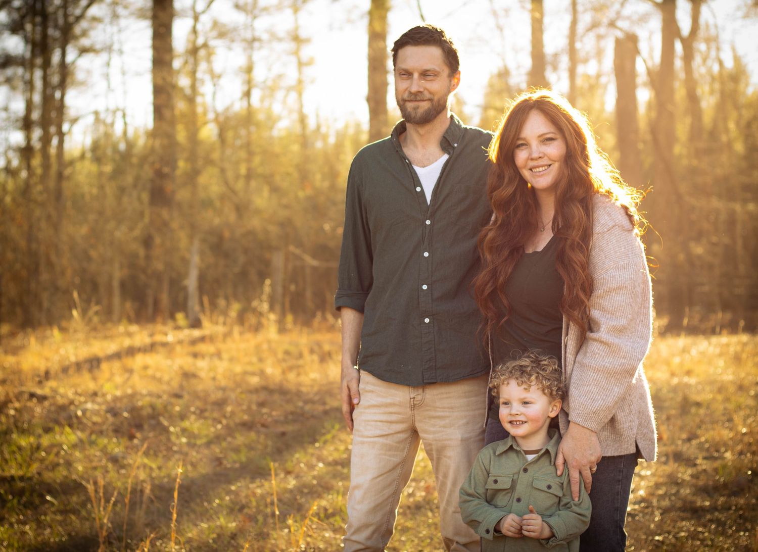 Family of three posing outdoors in golden sunlight among trees, parents smiling with young boy in green jacket.