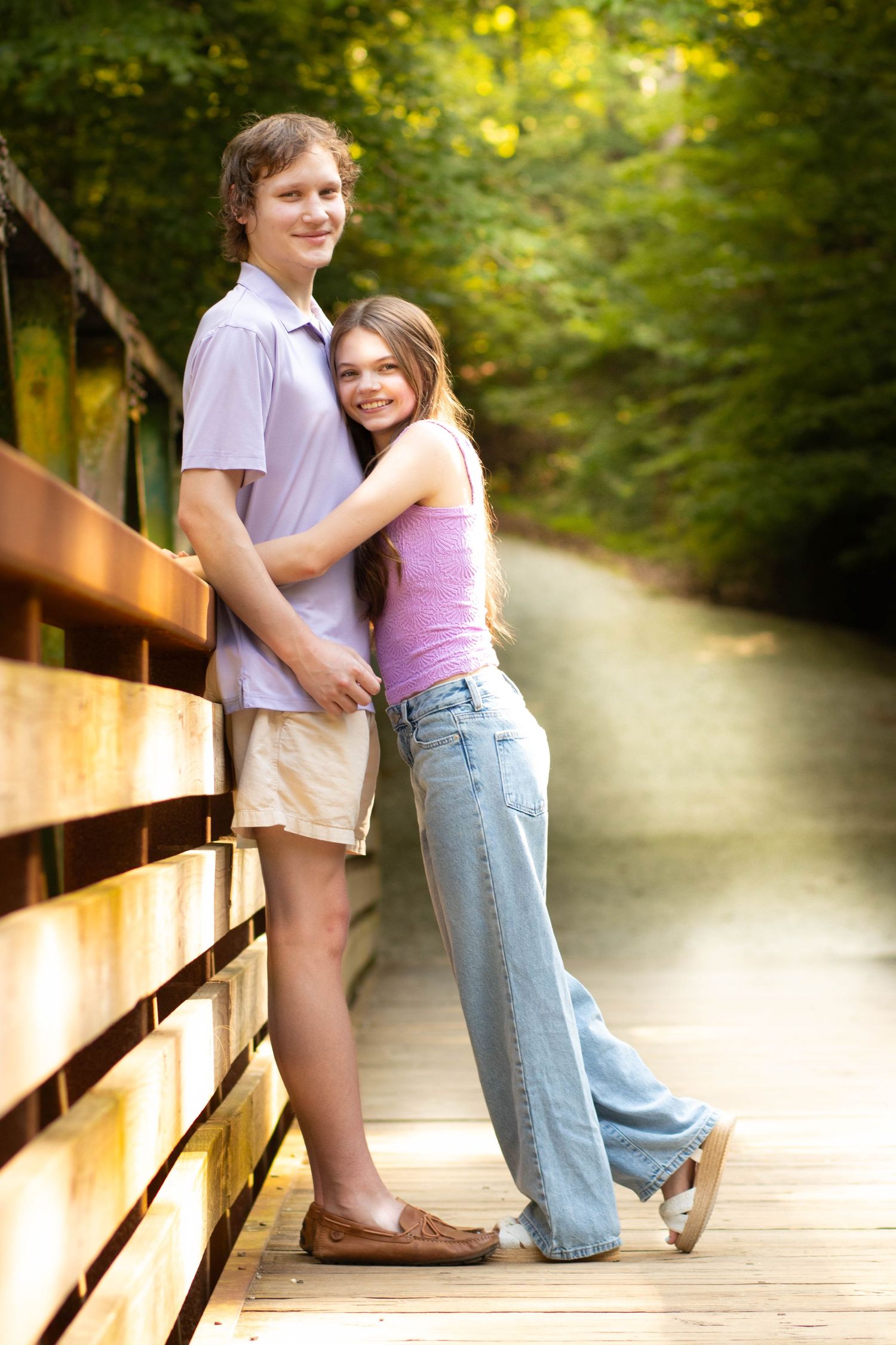 Young couple embracing on a wooden bridge surrounded by lush green trees in a romantic outdoor setting.
