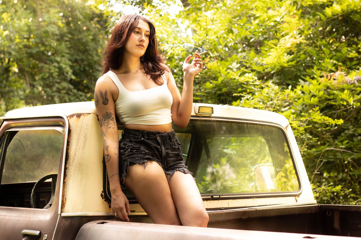 Young woman with tattoos in white crop top and shorts leaning on vintage pickup truck outdoors.