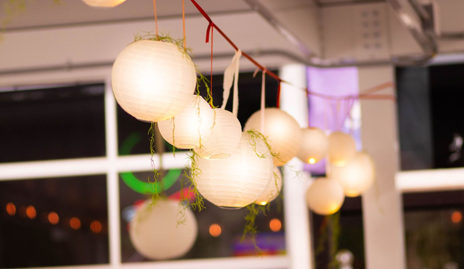 White paper lanterns hanging from red ribbon at an outdoor evening event with colorful bokeh lights in background.
