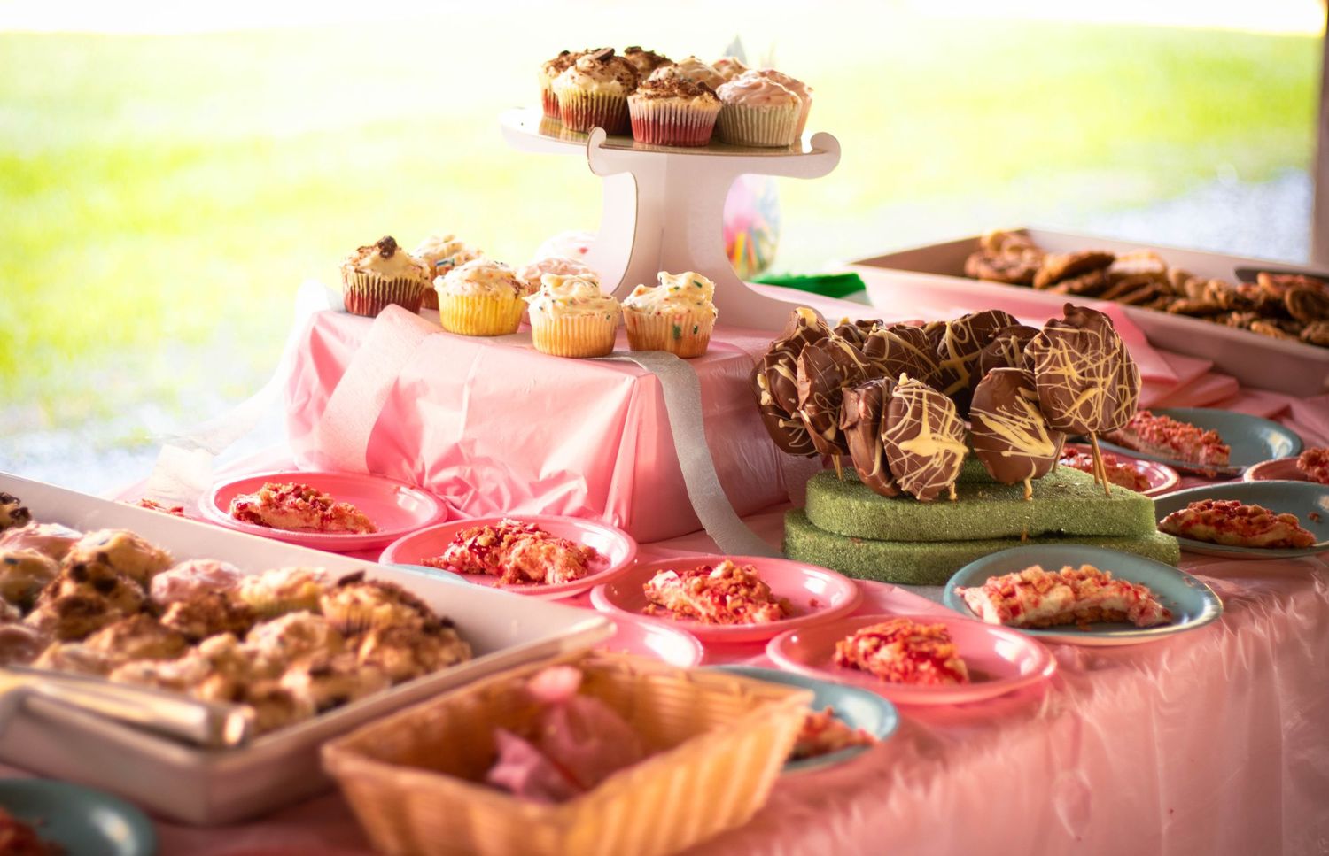 A pink-draped dessert table displaying muffins, brownies, and assorted sweets in colorful trays at an outdoor party.