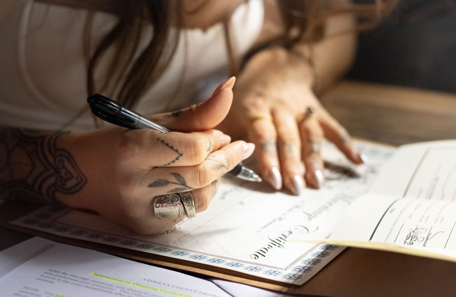 Tattooed woman writing on official documents with a pen at a wooden desk.