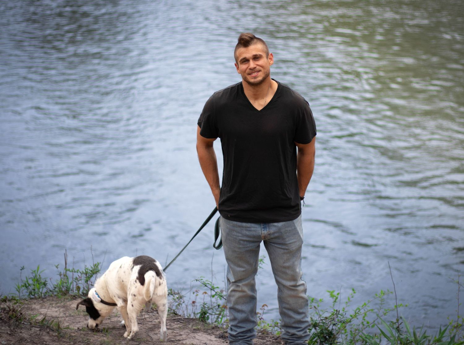 Man in black t-shirt walking white and black dog on leash near river bank on sunny day.