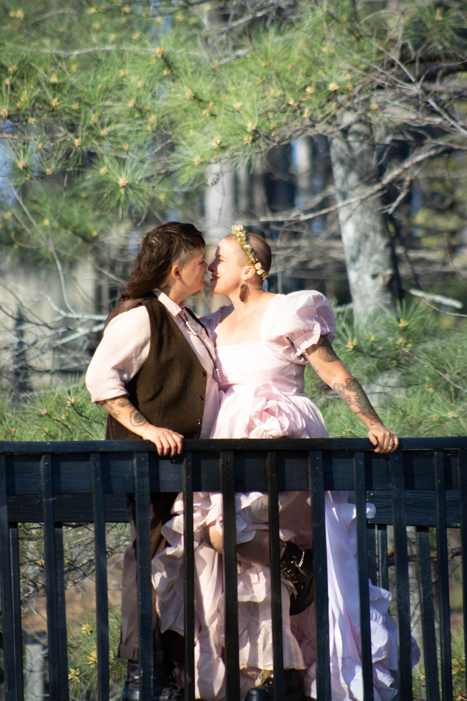 Couple in vintage attire sharing a romantic moment on a bridge surrounded by pine trees.
