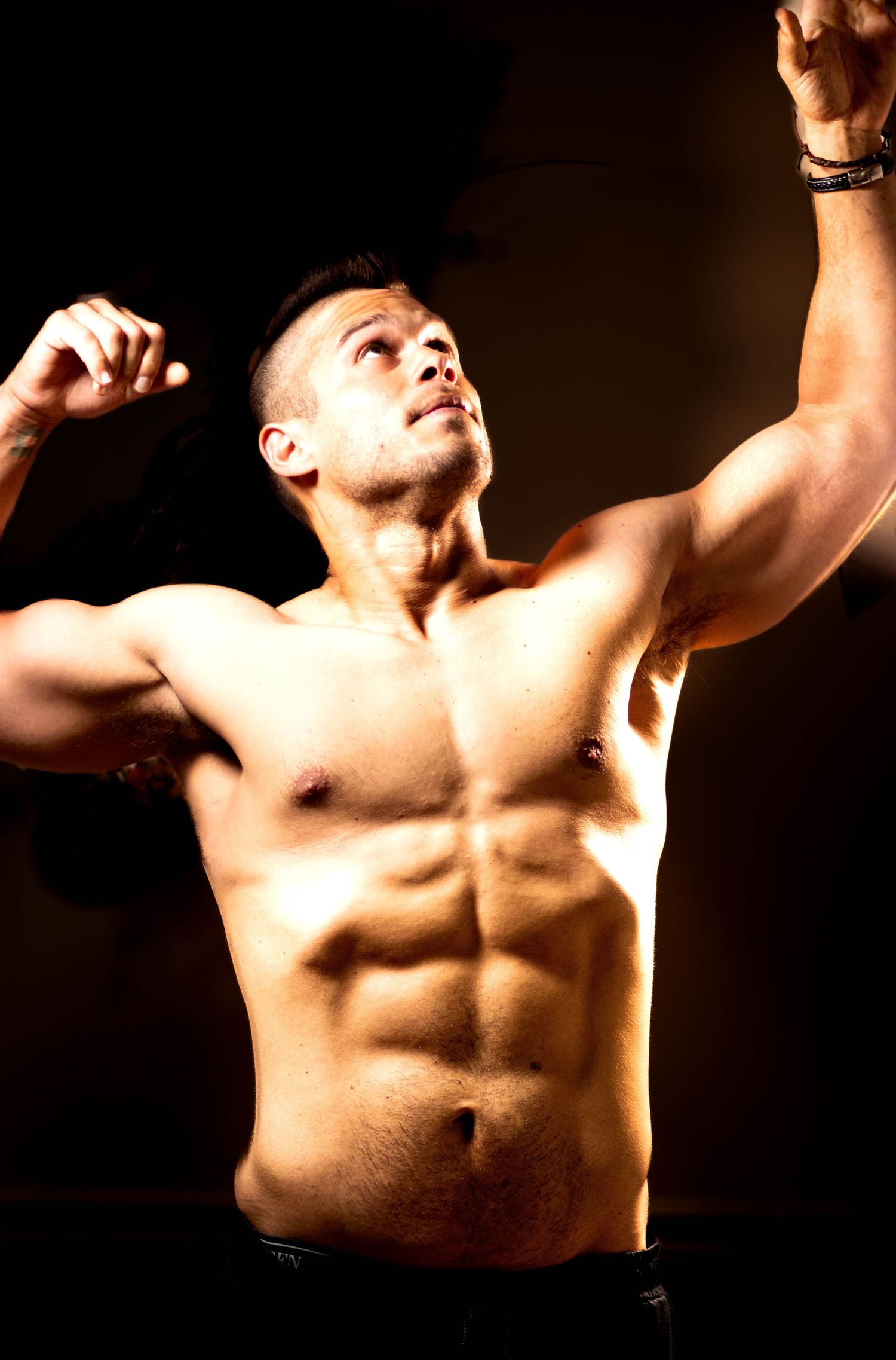 Muscular shirtless man flexing biceps with dramatic studio lighting highlighting defined abs and athletic physique.