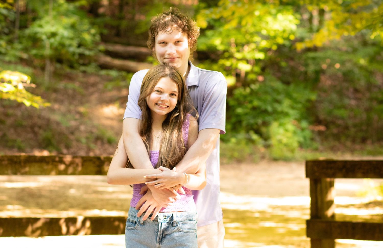 A young couple embracing and smiling outdoors on a wooden bridge surrounded by lush green trees.