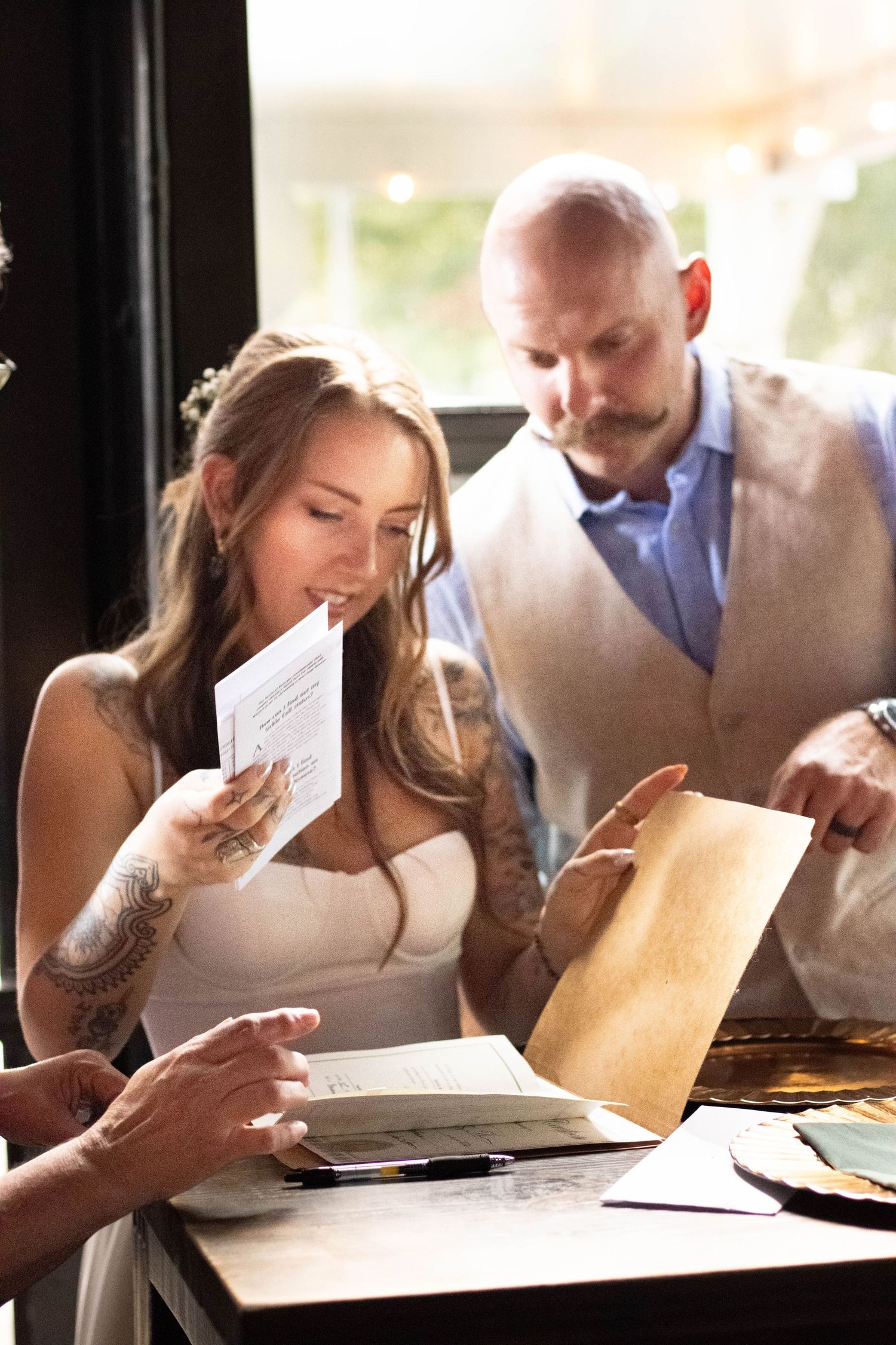Tattooed bride in white dress reads wedding card beside bald groom in vest at reception table.