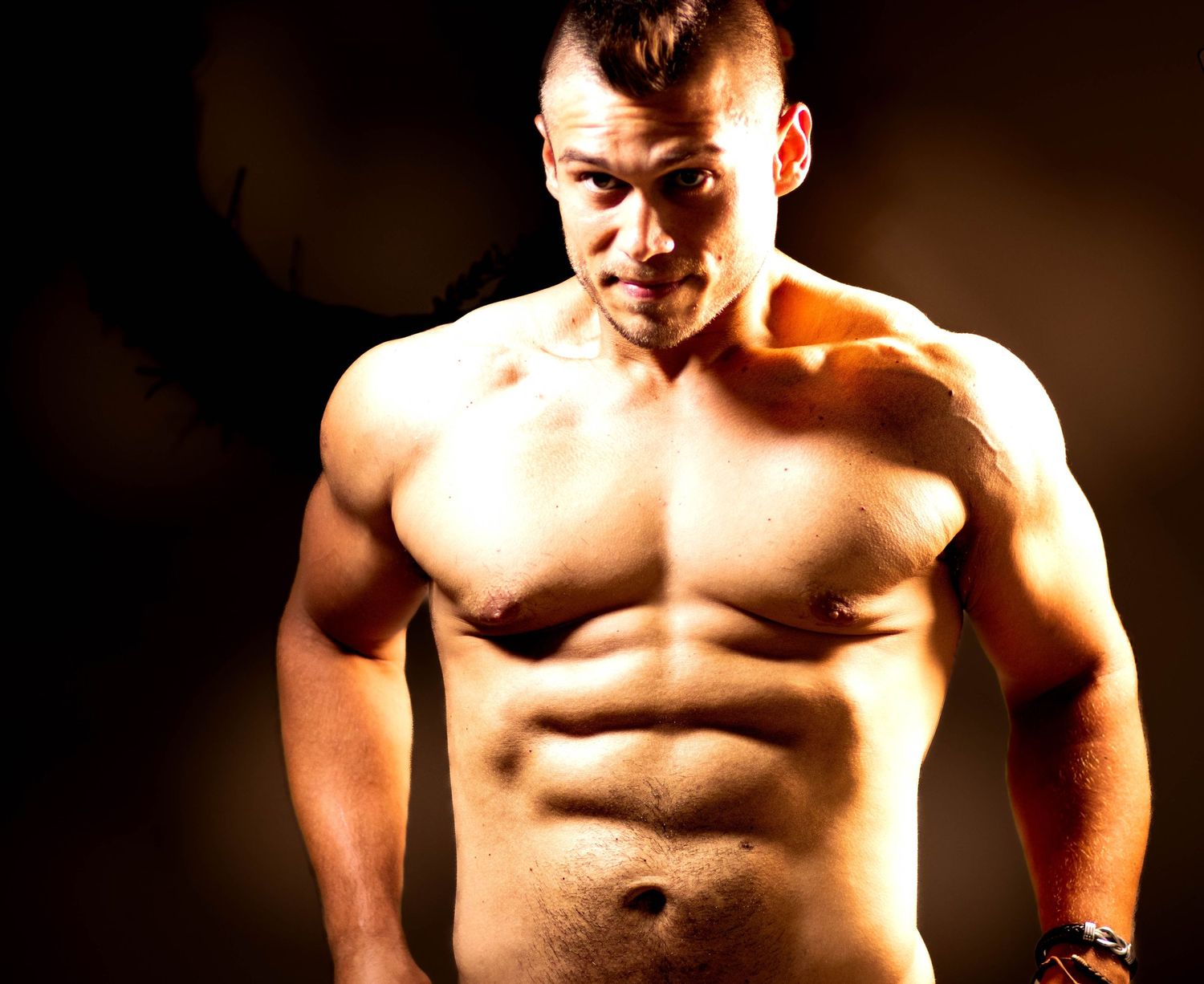 Muscular shirtless man with defined abs posing against dark background, showcasing athletic physique.