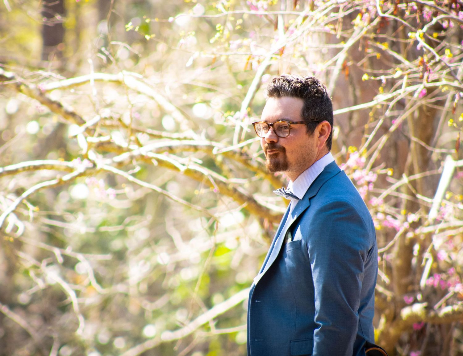 Elegant man in a blue suit with glasses standing among blooming branches at an outdoor event.