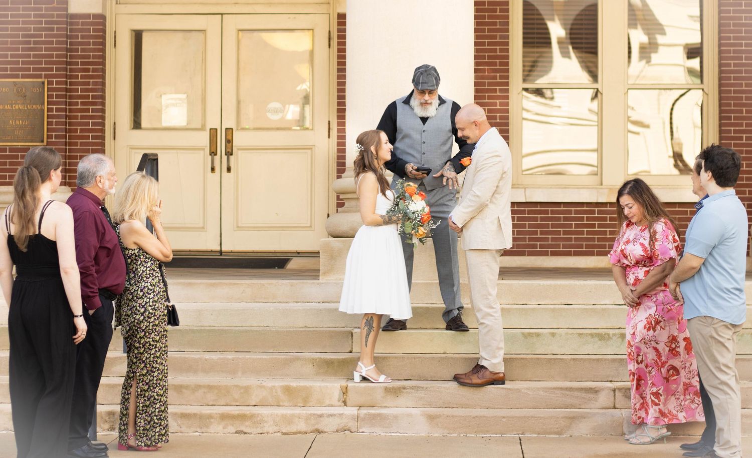 Outdoor wedding ceremony on church steps with bride holding bouquet, groom, officiant, and guests in warm evening light.