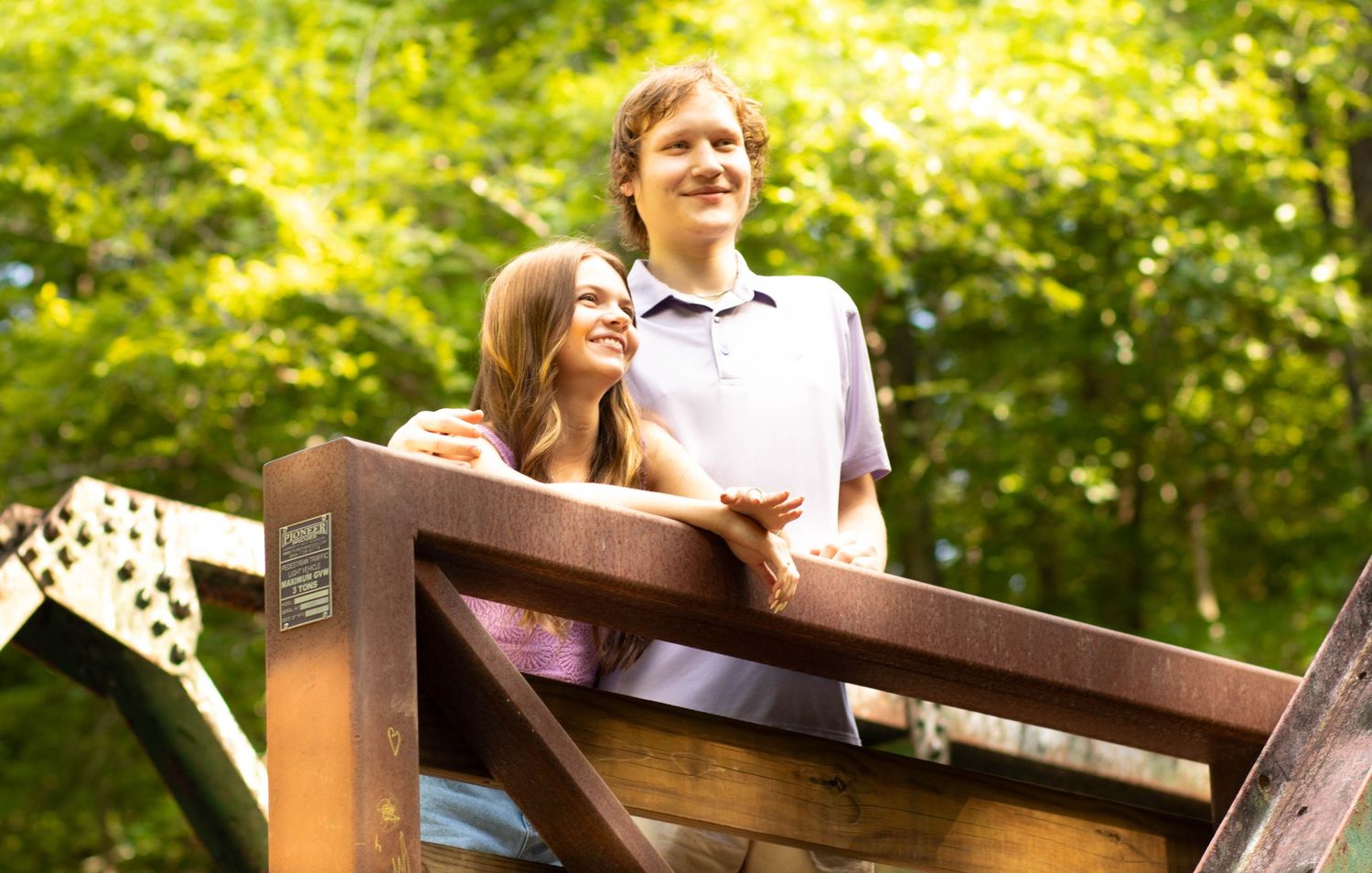 Two young people smiling and leaning on a wooden railing outdoors, surrounded by lush green trees.