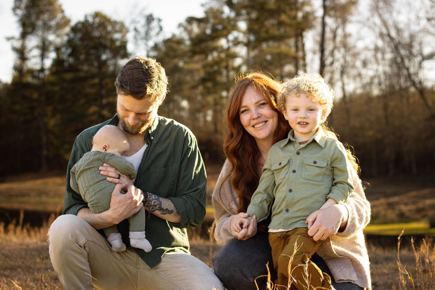 Family portrait outdoors with man holding baby, woman carrying toddler boy, smiling in golden field.
