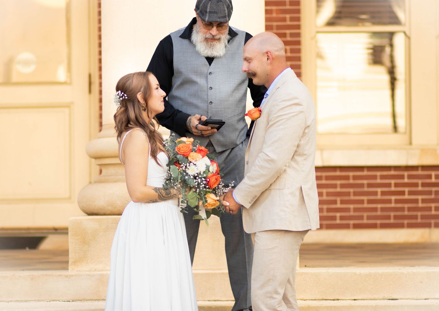 Bride and bald groom hold hands during outdoor wedding ceremony officiated by bearded man in gray vest.