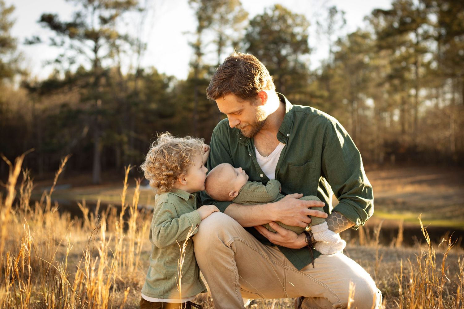 Father kneeling in sunny field tenderly holding newborn baby while young curly-haired toddler watches.