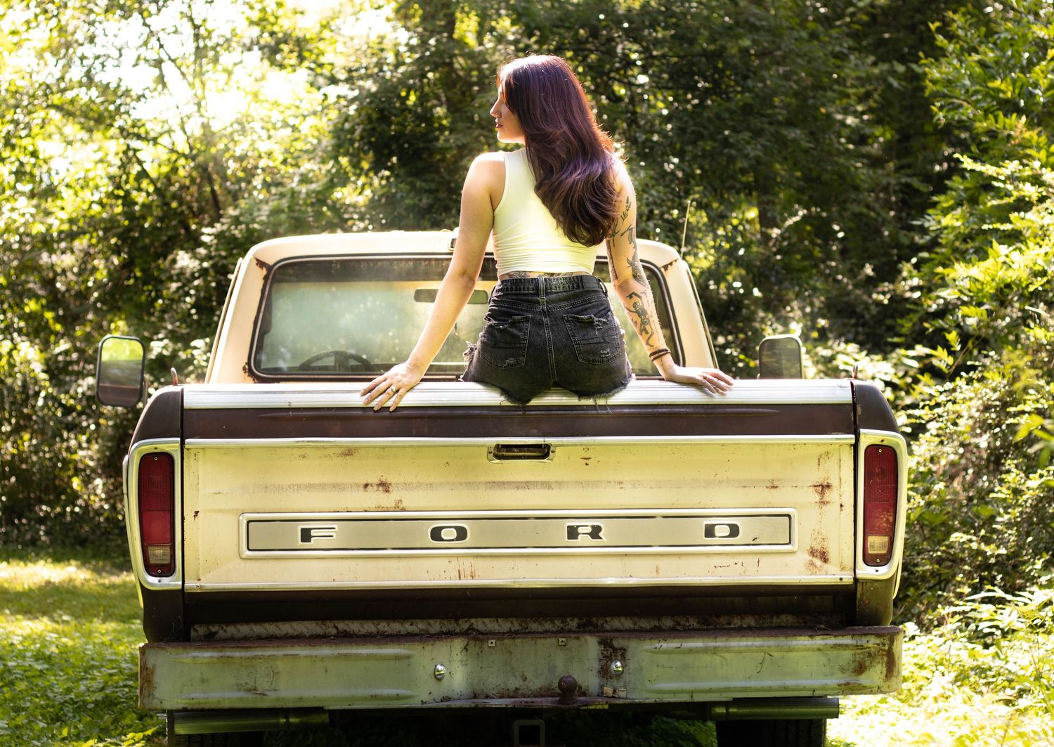 Young woman with purple hair sitting on the tailgate of a vintage Ford pickup truck surrounded by lush green trees.