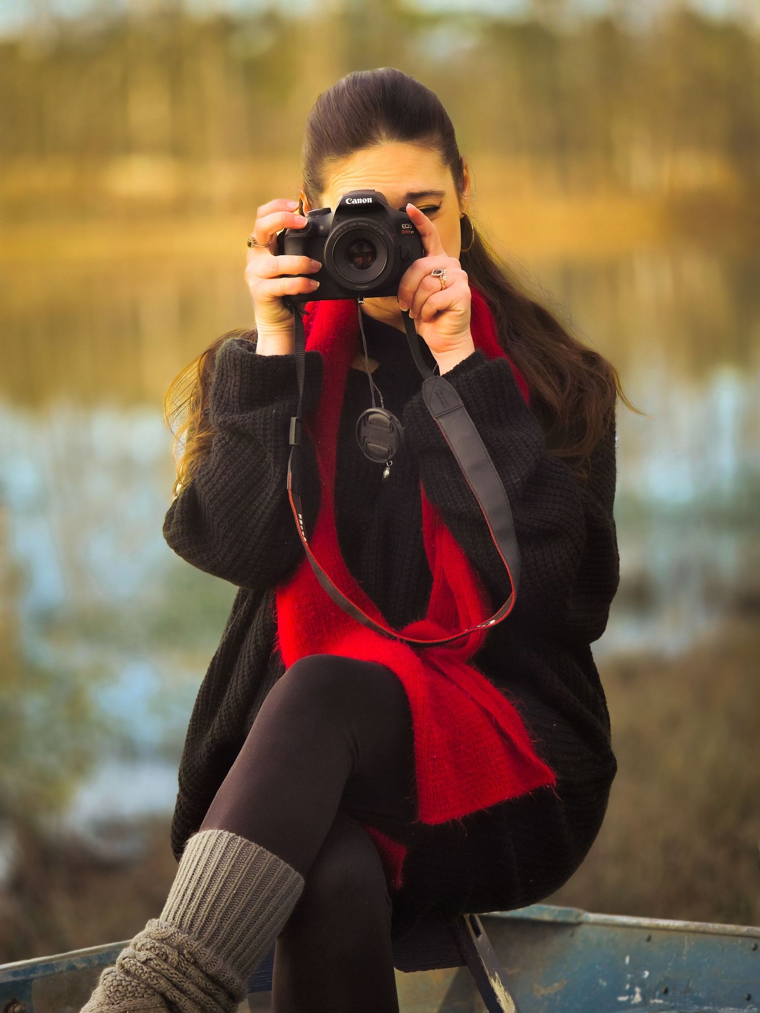 Female photographer aiming a Canon DSLR camera while seated outdoors near a lake.