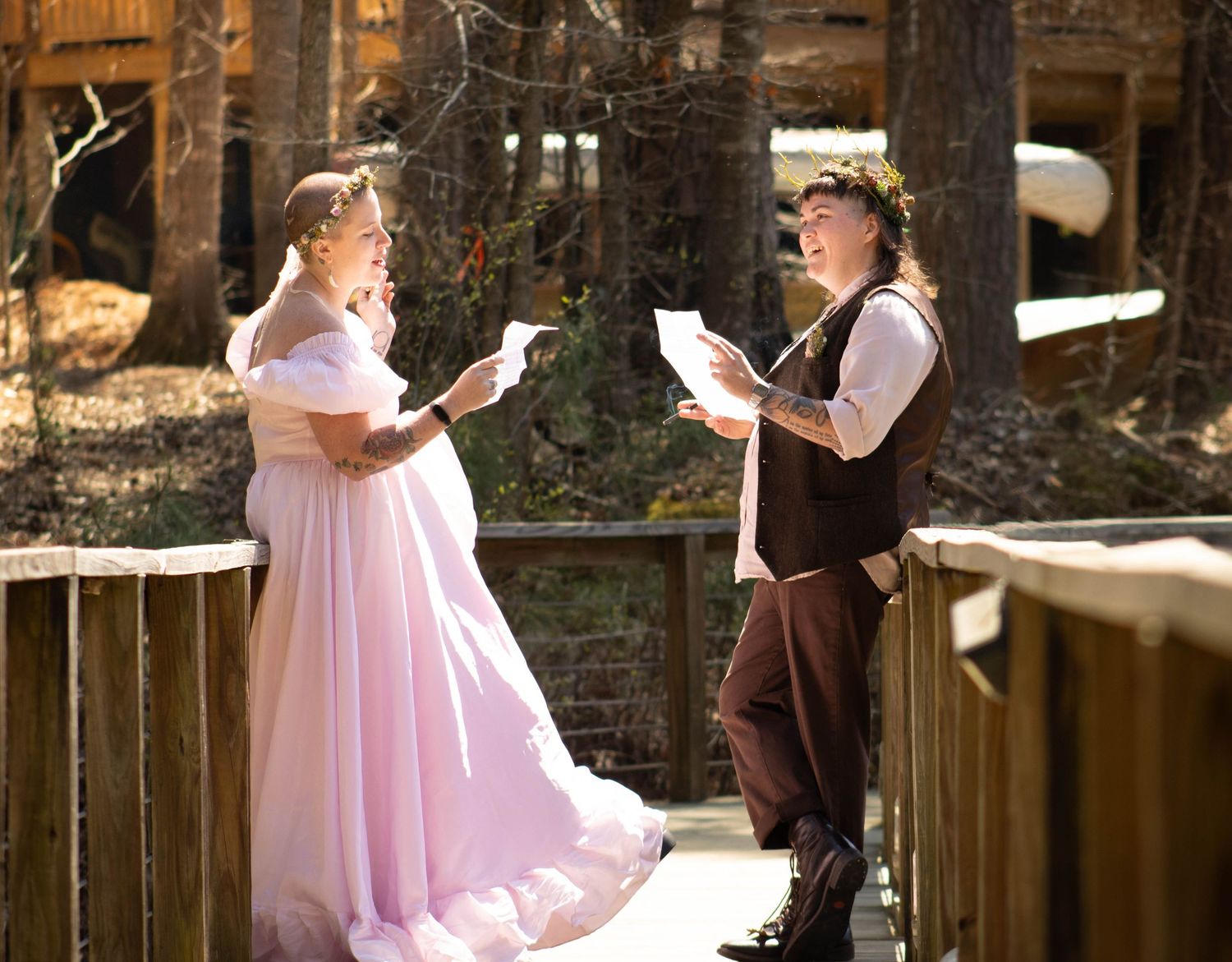 Couple in Renaissance costumes reading vows on a wooden bridge outdoors, woman in pink gown, man in brown vest.