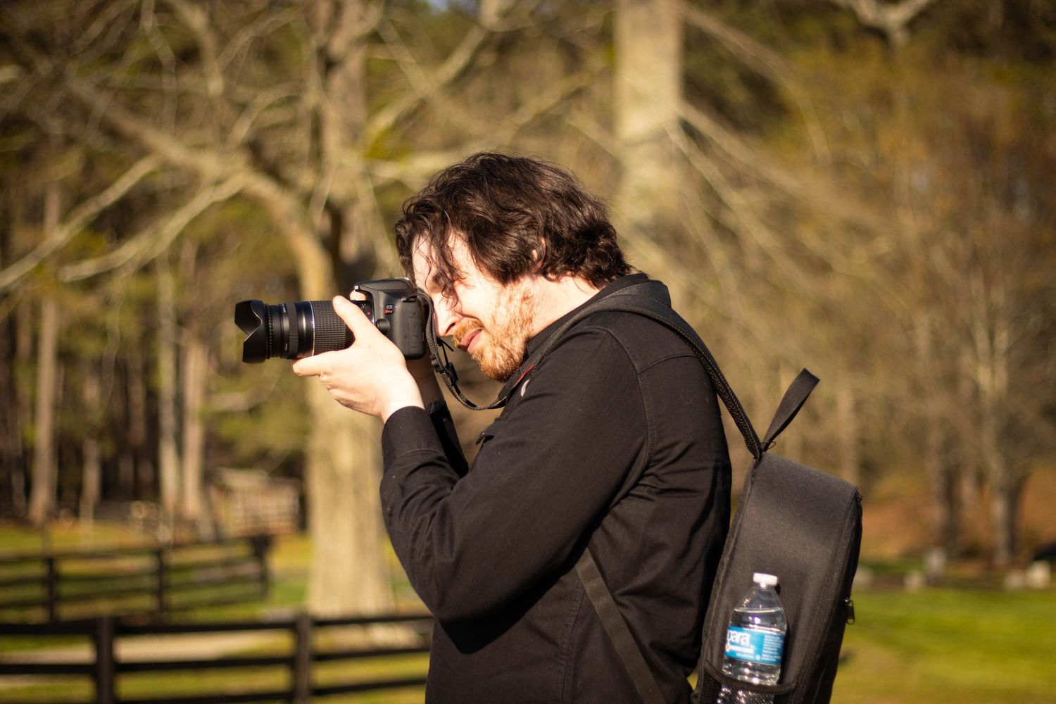 Male photographer with backpack shooting with DSLR camera outdoors in wooded park setting.