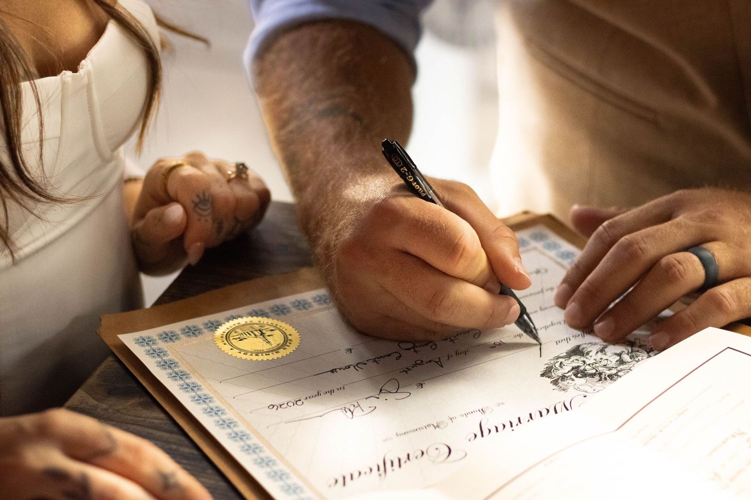 Close-up of hands signing a marriage certificate with a pen, with a gold seal visible on the document.