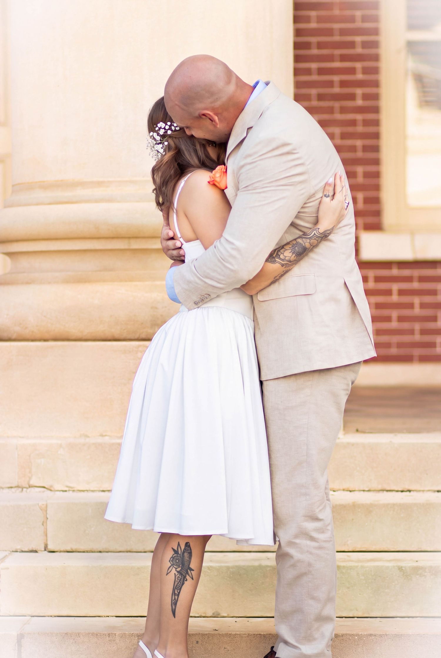 Couple sharing a romantic kiss on stone steps, bride in white midi dress with floral hair accessory and groom in beige suit.