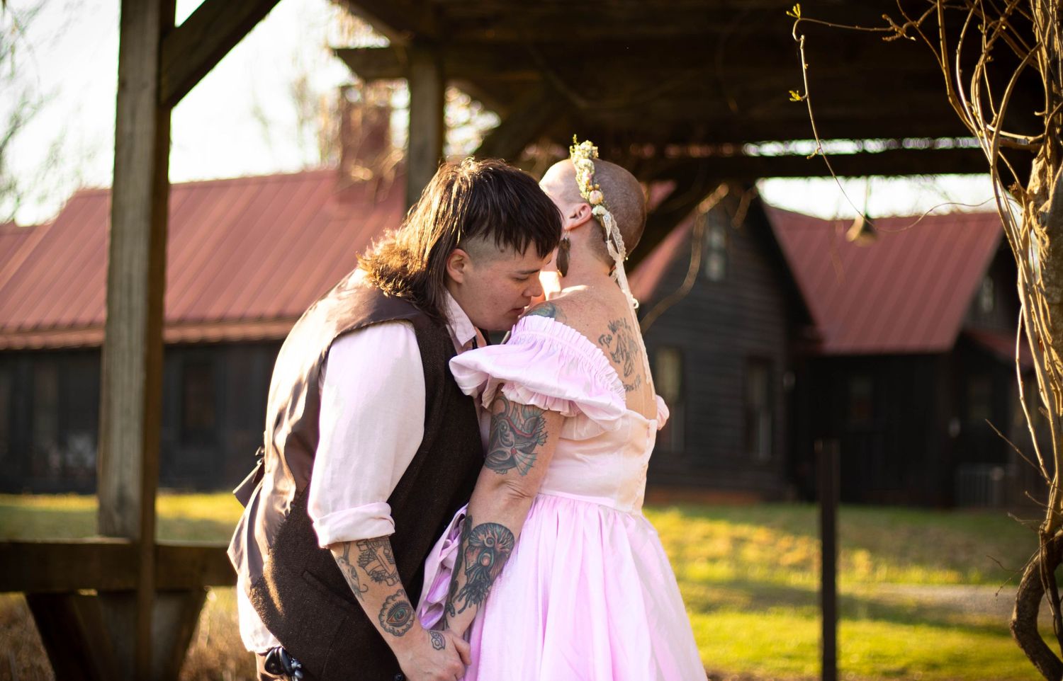Tattooed couple sharing a romantic moment in pink wedding attire outdoors near a rustic barn.