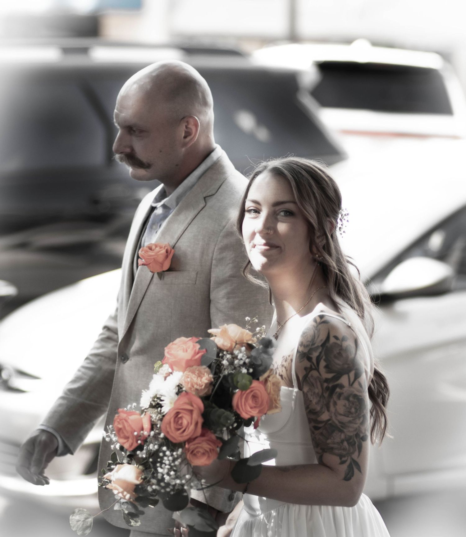 A bride with tattoos holding coral roses stands beside a bald groom in a gray suit with a matching boutonniere.