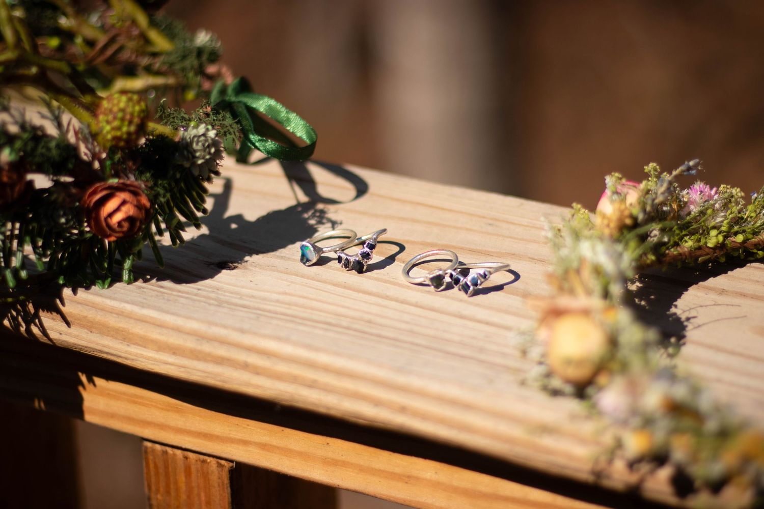 Two elegant rings with gemstones displayed on a wooden surface surrounded by floral wreaths in natural light.