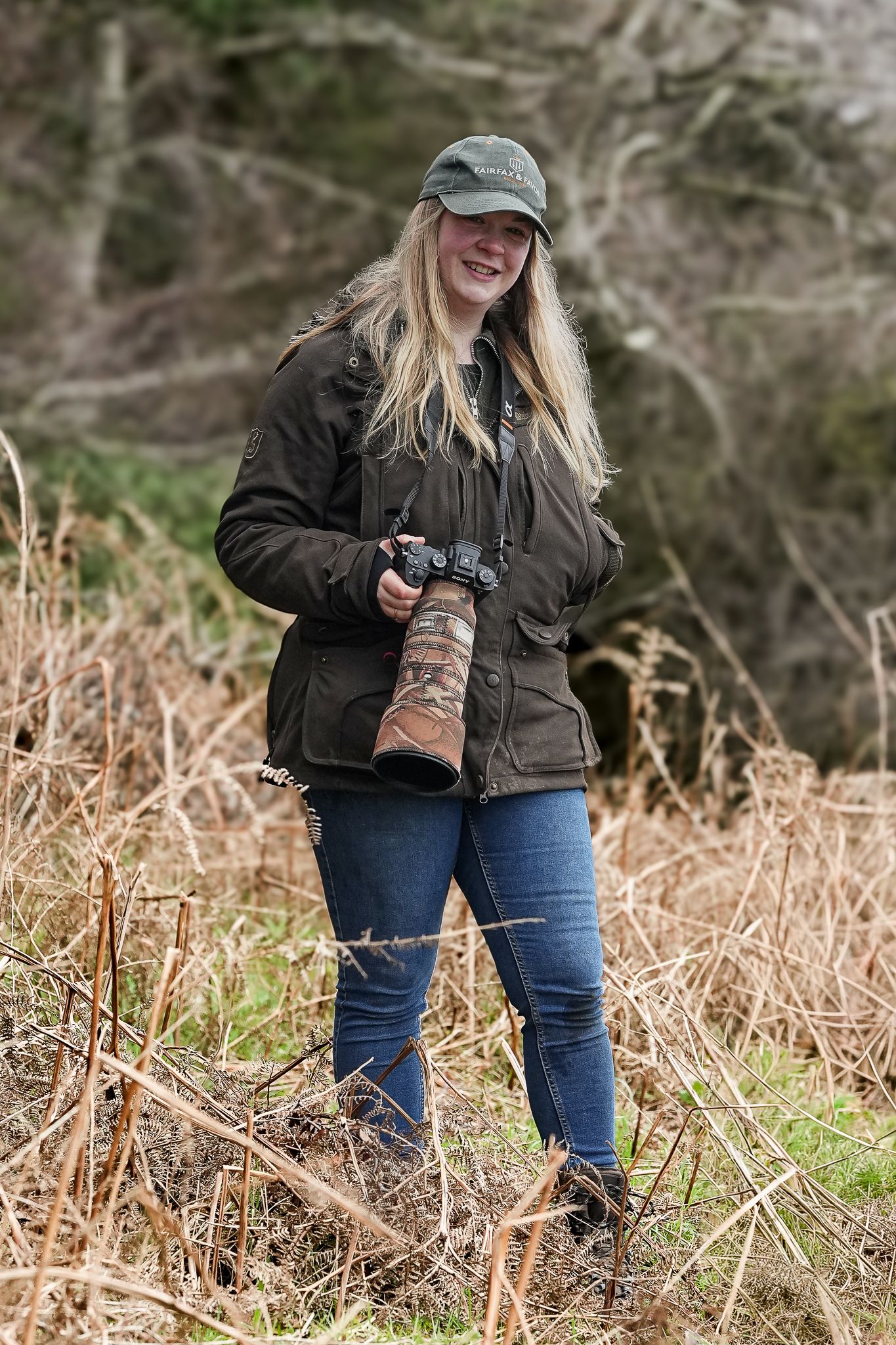 Smiling blonde woman in green cap holding professional camera with telephoto lens in outdoor setting.