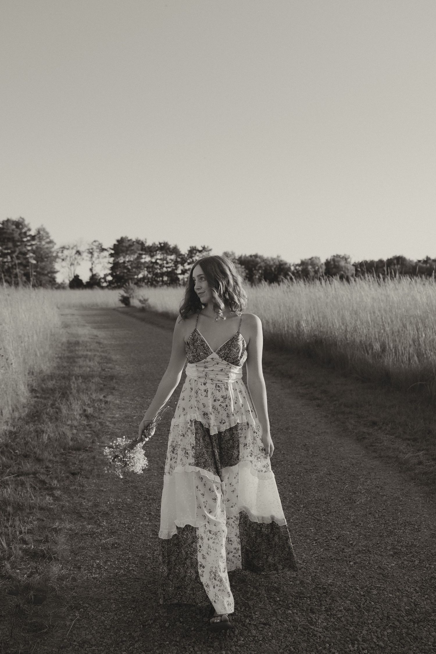 Black and white image of woman in floral dress walking along rural path holding wildflowers.