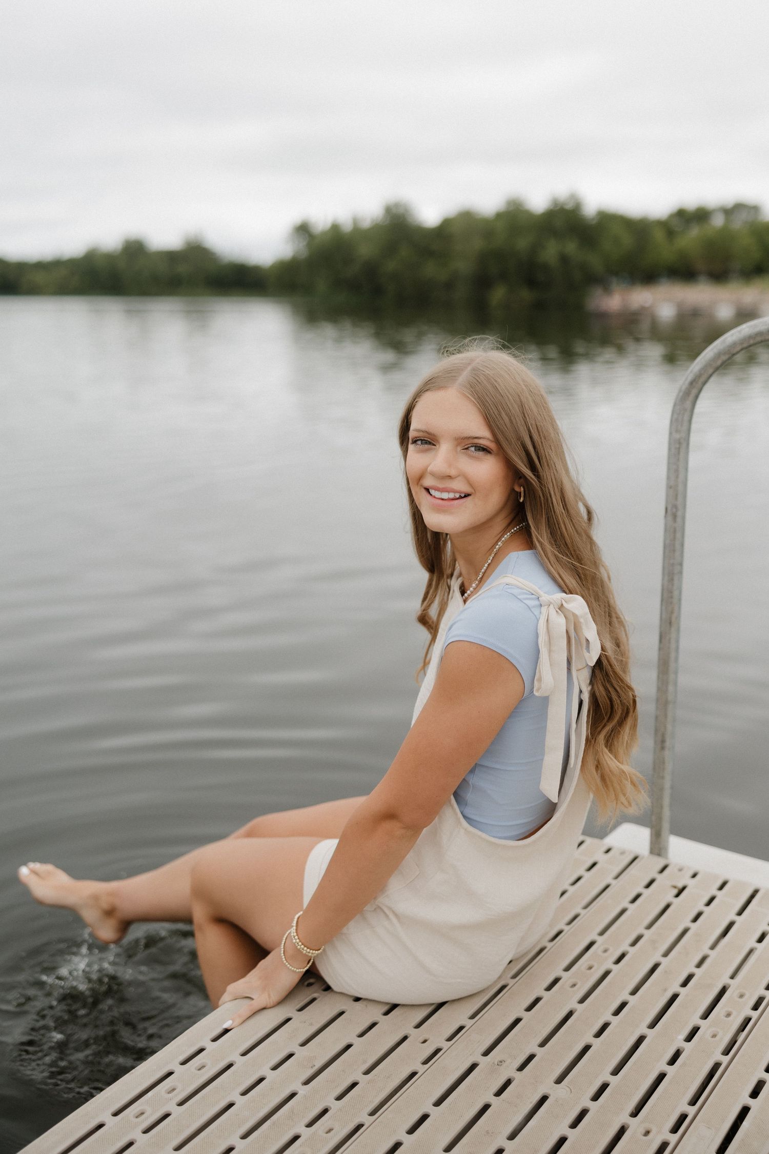 Smiling teenage girl in blue top and beige skirt sitting barefoot on a lake dock.