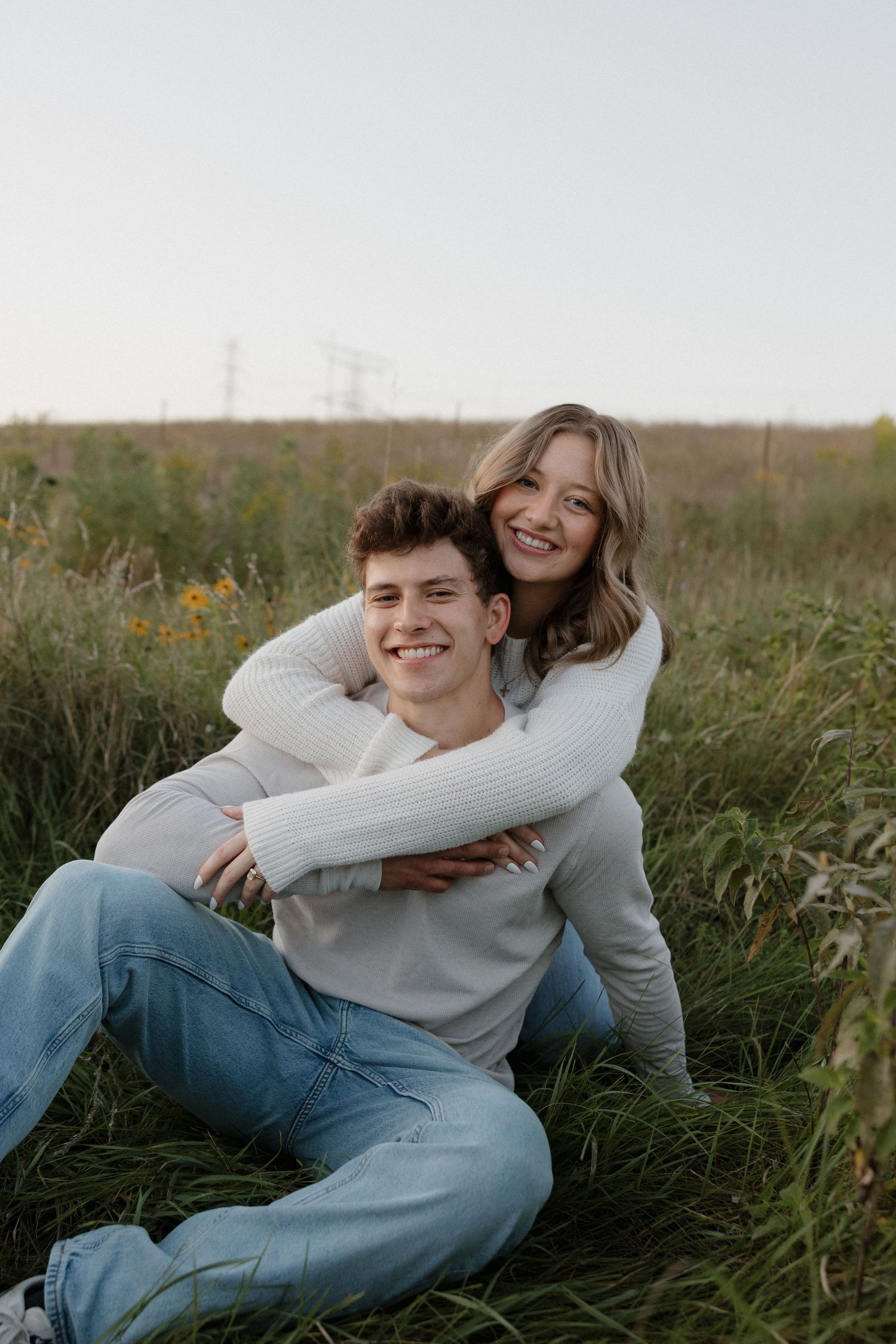Smiling couple sitting in a grassy field, woman hugging man from behind, both wearing matching cream sweaters.